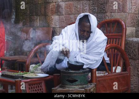 Lalibela, Äthiopien - November 2018: Mann in traditioneller weißer äthiopischer Kleidung, der Kaffeekörner röstet, bevor er einen Kaffee zubereitet Stockfoto