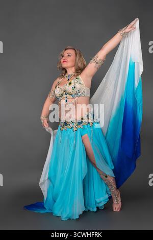 Lächelnde Bauchtänzerin mit Hand erhöht in Tanzbühnen-Outfit - BH-Oberteil und blauer langer Rock mit Schlitz und Pareo, der barfuß in voller Länge bewegt Stockfoto