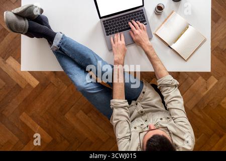 Blick von oben auf einen Mann in Smart, der an einem Laptop mit Füßen in Hausschuhen auf dem Schreibtisch arbeitet. Entspannte schlechte Haltung. Weißer Schreibtisch mit Notebook und Holzfußboden. Stockfoto