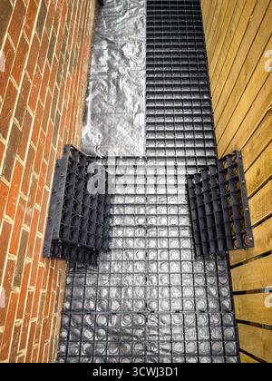 Schmaler Außenbereich mit schwarzen Abflussgittern zwischen Ziegelmauer und Holzzaun Stockfoto