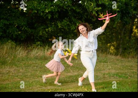 Mädchen und Mutter spielen glücklich mit großen Spielzeugflugzeugen auf dem Feld, jagen einander freudig. Mom und Tochter spielen draußen während des Urlaubs zusammen Stockfoto