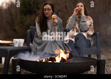 Zwei Frauen mit Decken sitzen draußen am Lagerfeuer und essen Marshmallows. Stockfoto