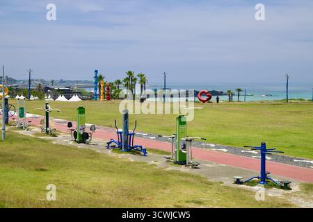Fitnessgeräte im Freien säumen einen Küstenpark oberhalb des Hamdeok Seoubong Beach mit öffentlichen Kunstwerken, Zelten und der blauen Bucht jenseits der Basaltmauern auf je Stockfoto