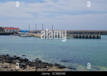 Der innere Kai am Hafen von Beollang zeigt kleine Fischerboote, die an einem Betonpier unter Versorgungsmasten befestigt sind, mit klarem Wasser über Sand und Basalt entlang der Stockfoto