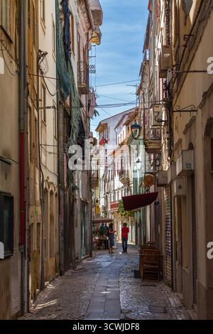 Eine enge, sonnendurchflutete Kopfsteinpflasterallee, die von historischen Gebäuden, Sitzgelegenheiten im Freien und hängenden Drähten unter einem klaren blauen Himmel gesäumt ist Stockfoto