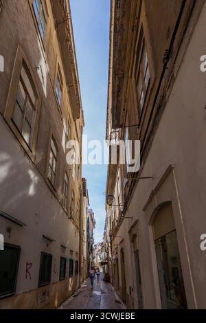 Eine schmale, sonnendurchflutete Gasse zwischen hohen, verwitterten Gebäuden umrahmt einen klaren blauen Himmel mit Fußgängern, die auf dem kopfsteingepflasterten Pfad darunter laufen Stockfoto