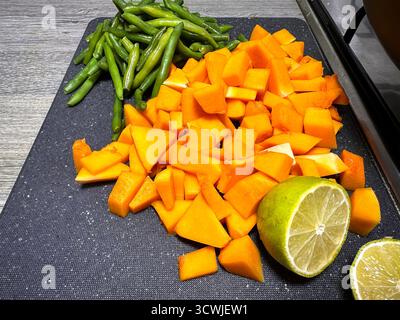 Gehackter Kürbis, frische grüne Bohnen und eine halbe Limette auf einem schwarzen Schneidebrett bereit zum Kochen Stockfoto