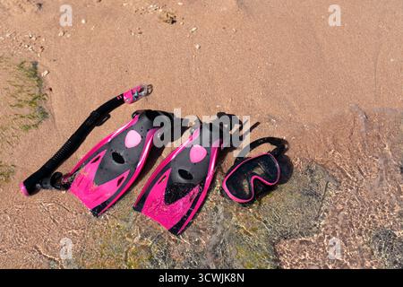 Ein Paar rosafarbene und schwarze Flossen, ein Schnorchel und eine Tauchermaske am Sandstrand und in der Nähe des flachen Wassers. Stockfoto