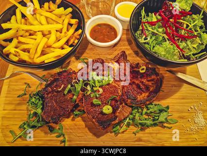 Close-up herzhafter Essensteller mit Steakkotelett auf einem Holzbrett über Rucola, garniert mit Schnittlauch und flankiert von Schüsseln mit goldenen Pommes Frites und gemischtem Salat. Sma Stockfoto