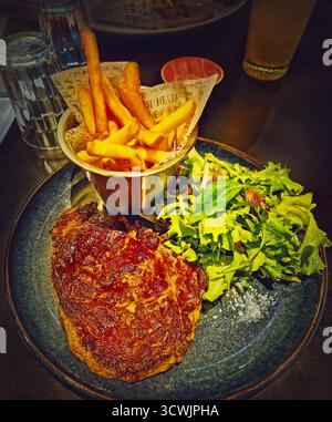 Rustikaler Teller mit einem großen, gut gebratenen Ribeye-Steak, serviert mit knusprigen goldenen Pommes frites und einem Haufen frischem grünem Salat Stockfoto
