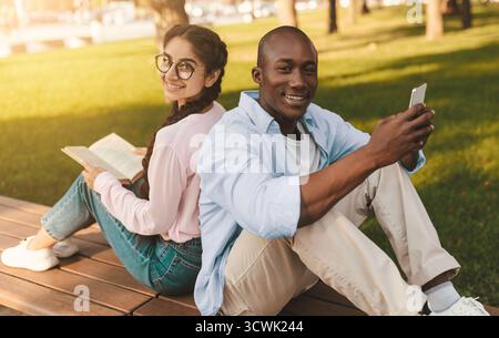 Glückliche, vielfältige Studenten, die sich mit dem Smartphone und dem Buch zurücklehnen und sich im Freien auf der Bank im Park ausruhen Stockfoto