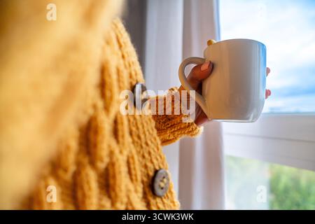Frau in einem gemütlichen gelben Pullover, der eine warme Tasse hält und sich am Fenster entspannt Stockfoto