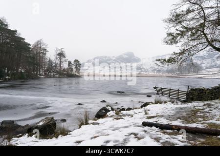 Gefrorenes Blea Tarn mit schneebedeckten Fjällen und Holzzaun in der winterlichen Landschaft des Lake District Stockfoto