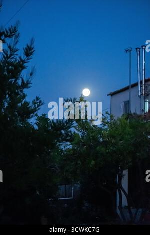 Der Vollmond leuchtet hell im blauen Abendhimmel über einem ruhigen Wohnhaus in einem mediterranen Dorf, das durch die dunklen Silhouetten betrachtet wird Stockfoto