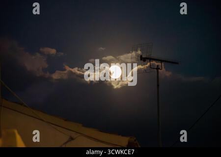 Der Vollmond scheint wie ein himmlisches Auge durch eine Pause in den dunklen Nachtwolken, mit einer Dachantenne, die sich vor dem geheimnisvollen und stimmungsvollen Licht abhebt Stockfoto