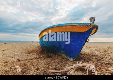 Altes Fischerboot ruht an einem Sandstrand unter einem dramatisch bewölkten Himmel. Die verwitterte Farbe und das rustikale Erscheinungsbild erinnern an Themen der Tradition, maritimen l Stockfoto