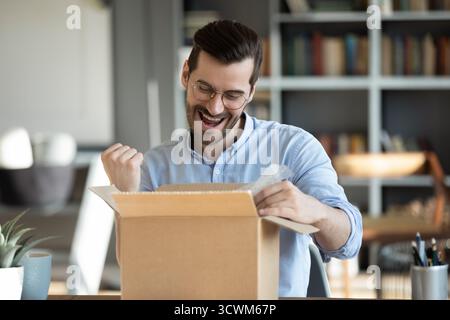 Ein glücklicher junger Mann, der das Postpaket am Schreibtisch öffnet Stockfoto