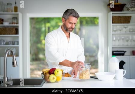 Mann isst morgens in der Küche. Der Mann genießt eine gesunde Mahlzeit zum Frühstück. Der Kerl isst Müsli und Obst zum Frühstück in der Küche Stockfoto