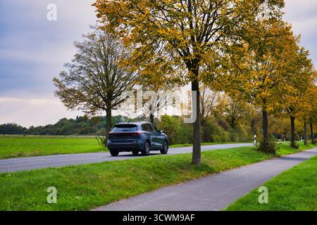 Dunkle Geländewagen fahren entlang der Rural Road, flankiert von lebhaften Bäumen des Goldenen Herbstes und grünen Feldern unter bewölktem Himmel in der Landschaft Stockfoto