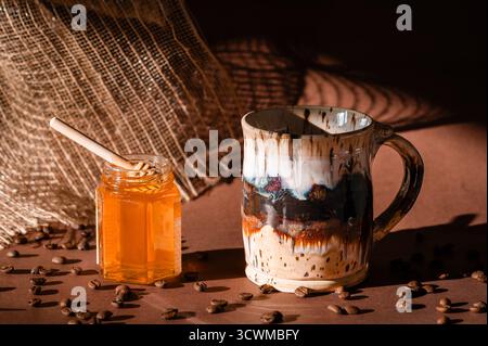 Eine Keramikkkaffeetasse und ein Glas Honig auf braunem Hintergrund, Kaffeebohnen im Vordergrund Stockfoto