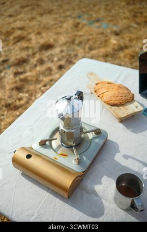 Kaffeekanne auf tragbarem Herd mit Brot und Metallbecher auf dem Tisch. Stockfoto