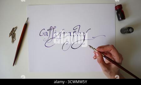 Handschriftliche Kalligraphie mit Tinte auf weißem Papier mit Tauchstift und Tintenflaschen. Hand einer Kaukasierin. Kunst, Kreativität, traditionelles Schreiben, handwerkliches Können und elegantes Design. Stockfoto