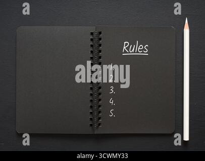 Öffnen Sie ein schwarzes Notizbuch, das neben einem weißen Bleistift eine nummerierte Liste von Regeln anzeigt, die auf einem dunklen Hintergrund stehen und die Einhaltung und Organisation veranschaulichen Stockfoto