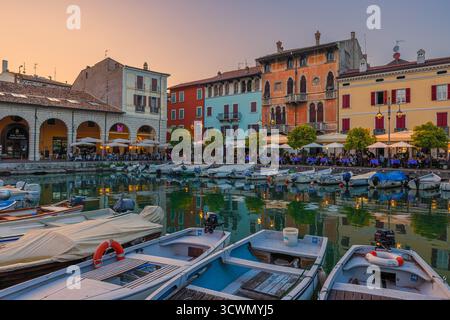 Lebendiger Sonnenuntergang über dem alten Hafen Porto Vecchio in Desenzano del Garda, Italien. Die belebten Terrassen und Restaurants am Wasser sind voll von Peop Stockfoto