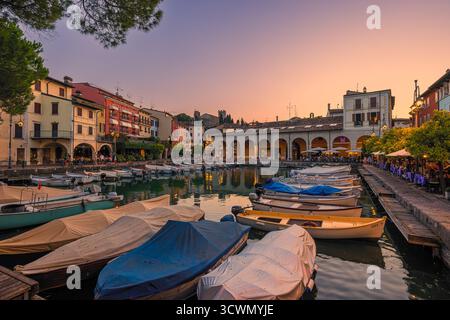 Lebendiger Sonnenuntergang über dem alten Hafen Porto Vecchio in Desenzano del Garda, Italien. Die belebten Terrassen und Restaurants am Wasser sind voll von Peop Stockfoto