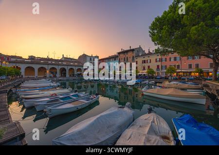 Lebendiger Sonnenuntergang über dem alten Hafen Porto Vecchio in Desenzano del Garda, Italien. Die belebten Terrassen und Restaurants am Wasser sind voll von Peop Stockfoto