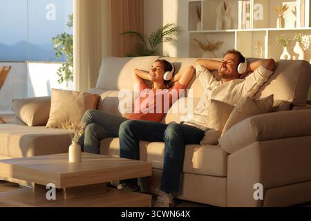 Glückliches Paar, das Kopfhörer trägt und Musik hört und zu Hause bei Sonnenuntergang auf einer Couch sitzt Stockfoto