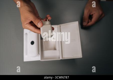 Draufsicht des jungen Mannes, der das weiße Smartphone-Ladegerät auspackt, das aus dem Karton ausgepackt wird, auf einem grauen Tisch sitzt. Stockfoto