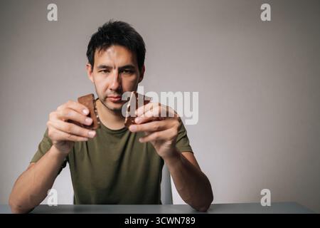 Porträt eines jungen Mannes, der an einem Tisch sitzt und die Schokoladentafel in zwei Stücke zerbricht, während er darüber nachdenkt, welches Stück zuerst gönnen soll Stockfoto