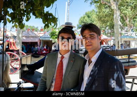 Sevilla, Spanien - 23. April: Zwei junge Männer, die bei einer festlichen Veranstaltung im Freien posieren, in stilvollen Outfits, mit farbenfrohen Dekorationen und Menschen im bac Stockfoto