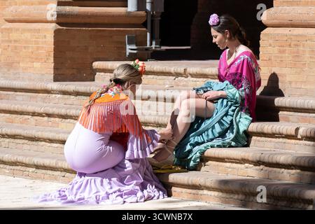 Sevilla, Spanien - 23. April: Zwei Frauen in traditionellen Kleidern sitzen auf Treppen, eine passt die Schuhe an, lebhafte Farben und kulturelle Kleidung Stockfoto