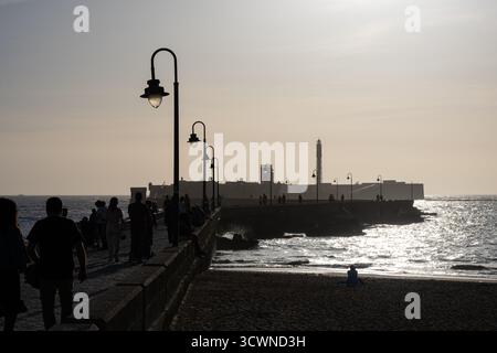 Silhouettenfiguren, die bei Sonnenuntergang am Pier von Cadiz, Spanien, spazieren, mit einem Leuchtturm im Hintergrund und sanften Wellen, die das Licht reflektieren Stockfoto