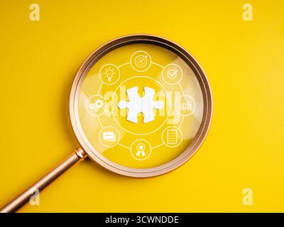 Lupe mit Schwerpunkt auf Puzzleteil mit Symbolen für die Geschäftsstrategie, die den Einstellungsprozess für Mitarbeiter, die Stellensuche, die Kandidatenauswahl und die Rekrutierung darstellen Stockfoto