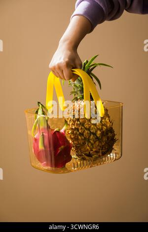 Hand mit einem Korb mit frischen tropischen Früchten, Ananas und Drachenfrüchten in einer Glasvase auf braunem Hintergrund Stockfoto