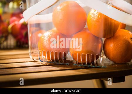 Frische Orangen in einem transparenten Korb auf einem Holztisch, Nahaufnahme von Orangen in einem Kunststoffkanister auf einem Regal Stockfoto
