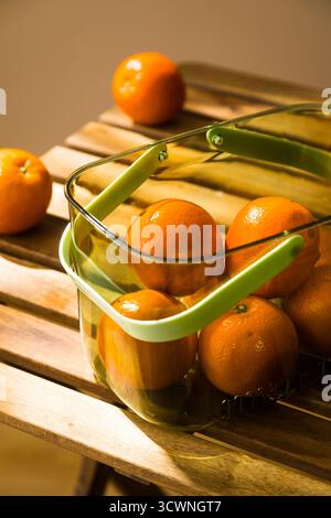 Frische Orangen in einem klaren Korb auf einem rustikalen Holztisch, frische Mandarinen in einem Metallkorb auf einem Holztisch. Stockfoto