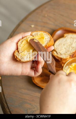 Goldene Butter auf ein frisches Stück Brot verteilen, Frauenhände schneiden Brot mit Butter auf einem Holzbrett, Nahaufnahme Stockfoto