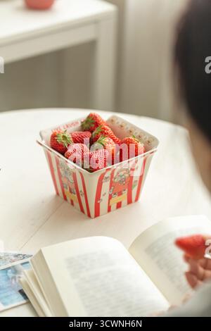 Genießen Sie frische Erdbeeren beim Lesen eines Buches, Mädchen beim Lesen eines Buches und essen Erdbeeren. Selektiver Fokus auf Erdbeere. Stockfoto