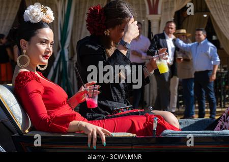 Sevilla, Spanien - 23. April 23: Zwei Frauen in traditioneller Kleidung sitzen in einer Kutsche und genießen bunte Getränke, mit einem festlichen Hintergrund von Menschen und d Stockfoto