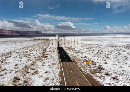 Aus der Vogelperspektive auf eine lange, dunkle Straße, die durch eine schneebedeckte Landschaft führt, im Kontrast zu den fernen roten Felsformationen unter einem hellen, teils bedeckten Felsen Stockfoto