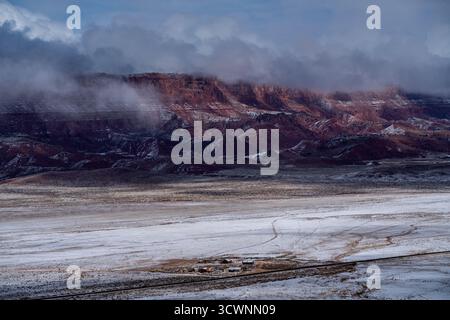 Aus der Vogelperspektive des schneebedeckten Wüstenbodens trifft auf die zerklüfteten roten Felsformationen, die in ätherischen Wolken gehüllt sind, und schafft einen starken Kontrast zwischen Texturen und t Stockfoto