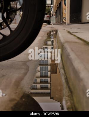 Motorradpfütze: Nahaufnahme eines Radreifens, der über einer Wasserreflexion auf einer grauen Stadtstraße schwebt und ein surreales, stimmungsvolles und gedämpftes Gefühl vermittelt. Stockfoto