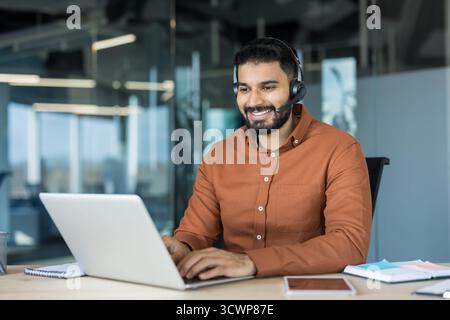 Lächelnder junger Mann, der am Schreibtisch arbeitet, ein Headset mit Mikrofon trägt und Online-Support in einem modernen Callcenter oder Home Office bietet Stockfoto