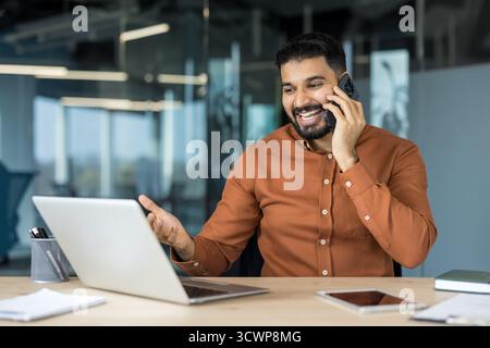 Ein indischer Geschäftsmann lacht und gespendet, während er auf einem Mobiltelefon an einem Schreibtisch mit einem Laptop sitzt und in einer hellen, modernen Büroumgebung ein lebhaftes Gespräch am Arbeitsplatz genießt Stockfoto