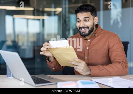 Ein aufgeregter junger professioneller Mann, der an seinem Schreibtisch sitzt und einen braunen Umschlag mit einem überraschenden, glücklichen Ausdruck öffnet und von einer Beförderung oder einem finanziellen Erfolg erfährt Stockfoto