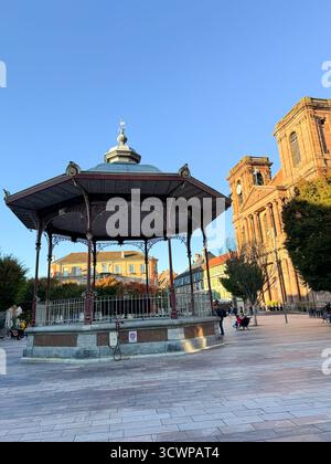 Verzierte gusseiserne Bühne auf dem zentralen Platz von Belfort mit historischer Kirche und Uhrenturm im Hintergrund unter der warmen Nachmittagssonne Stockfoto